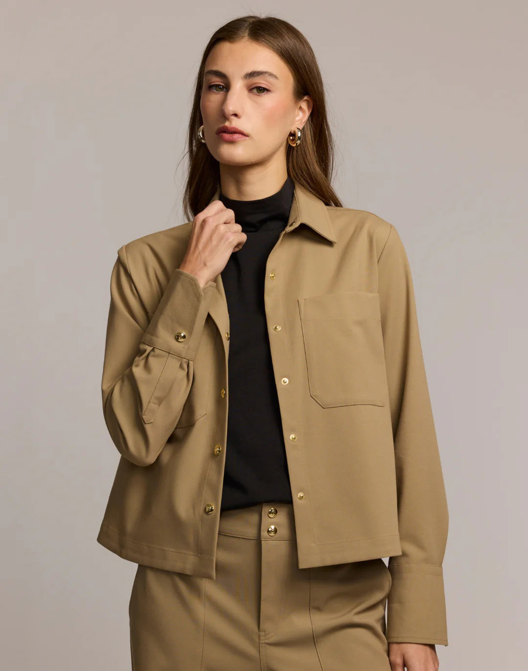 A woman with straight brown hair and gold hoop earrings is seen from the back in the HINSON WU ASTRID Long Sleeve Traveler&#39;s Twill Shirt Jacket, styled for travel, against a plain background.