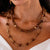 Close-up of a person wearing two necklaces. The shorter necklace, part of the CAPUCINE DE WULF - EARTH GODDESS SINGLE STRAND NECKLACE collection, has small circular wooden beads with a few textured gold beads interspersed, adding an element of earthy opulence. The longer necklace features larger hand-carved teak beads connected by 18K Gold IP over Brass loops. The person is wearing a white outfit.