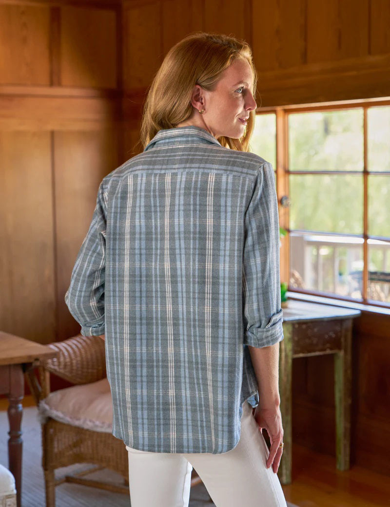 A person with long hair stands in a wooden room next to a window, wearing a relaxed fit FRANK & EILEEN EILEEN button-up shirt in grey cream blue plaid Italian flannel and white pants. Light floods the wicker chair and wooden walls.