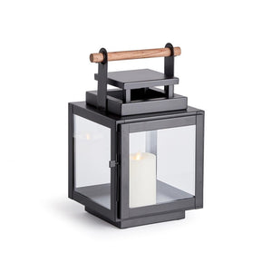 The ADWIN OUTDOOR LANTERN by NAPA HOME AND GARDEN is a black metal lantern with glass panels and a teak handle on top. Inside, there's a white pillar candle. The design is modern and exclusive, featuring clean lines and a sturdy structure, making it an ideal choice for both indoor and outdoor use.