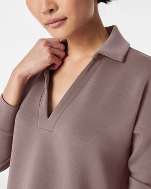 A person with dark hair styled in a neat updo is wearing the SPANX - AirEssentials Split Neck Polo Top in black, paired with matching black pants. They rest one hand on the back of their neck and gaze to the side against a plain white background, exuding an air of luxurious comfort.