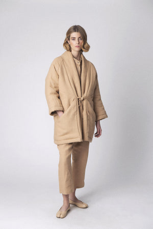 Against a light gray backdrop, a person with short hair models the LOESS MONICA Linen Puffer Coat in beige, complete with matching pants and light slippers. This minimalist outerwear, tied at the waist, captures the essence of transitional weather layering as they stand relaxed.