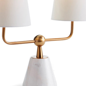 The MADISON DUBLET LAMP by NAPA HOME AND GARDEN showcases a sleek, contemporary design with a sophisticated white, conical marble base and two elegant white cylindrical lampshades. The lamp is supported by a gold metallic bar that gracefully splits into two upward arms featuring a brass finish, embodying modern elegance.