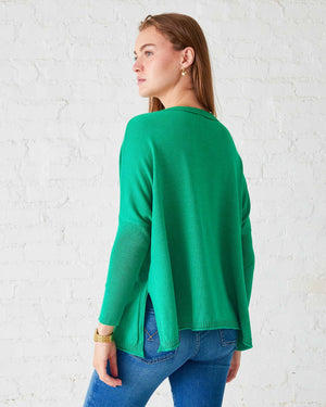 A woman in a versatile green MERSEA - CHEERS CATALINA CREWNECK SWEATER, featuring the word "cheers," stands against a white brick wall, smiling with her hands at her sides. This staple piece from MER SEA pairs perfectly with blue jeans and a gold bracelet.