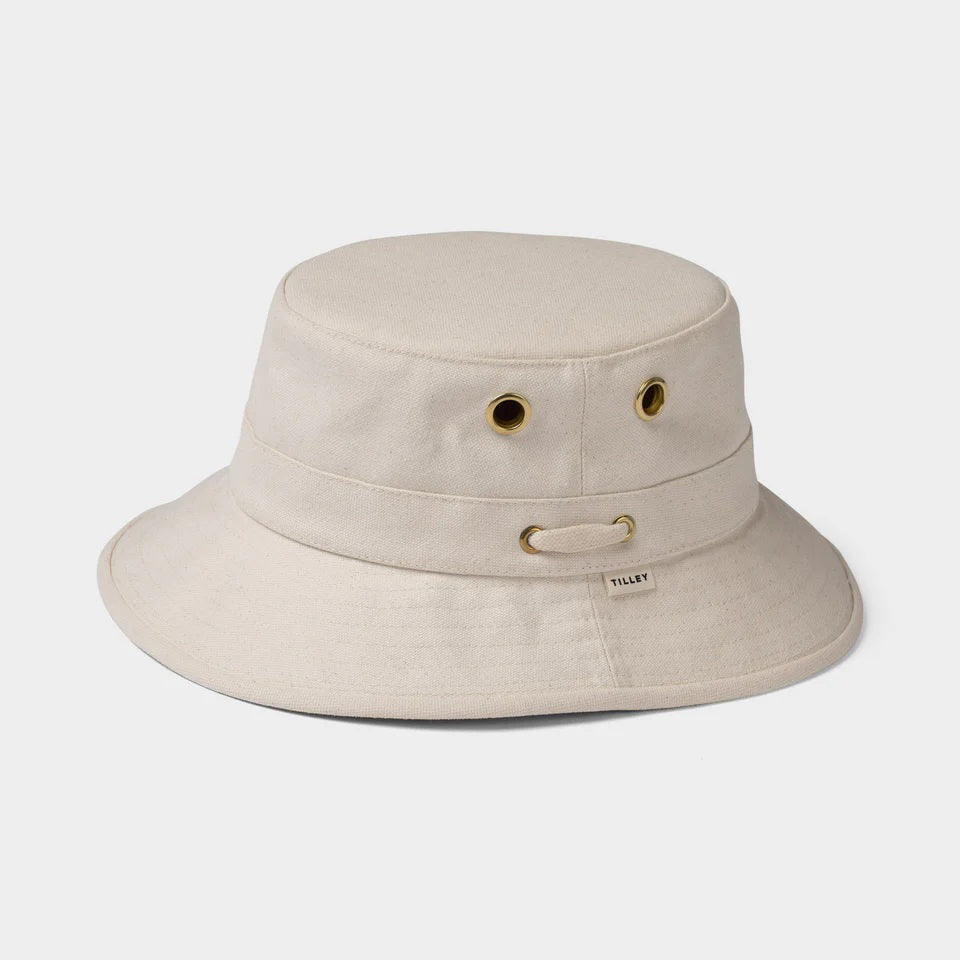 The TILLEY - ICONIC T1 BUCKET HAT by TILLEY is a beige cotton sun hat with a wide brim, two gold side eyelets, a small fabric tag, and chin strap loop, shown against a plain light grey background.