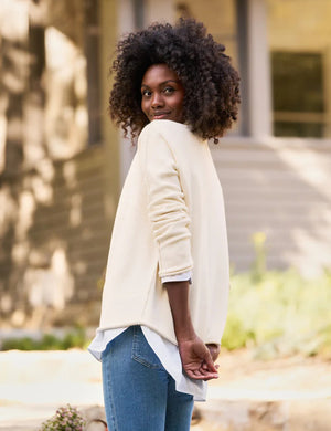 A woman with curly hair is wearing the luxurious FRANK & EILEEN - MONTEREY Rolled Funnel Neck Sweater in cream over a white collared shirt. She is looking to her right, and the background features outdoor greenery with a soft focus.