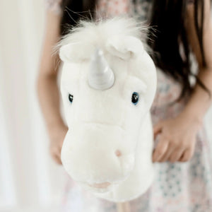 A young child in a cozy, bohemian-style bedroom holds the LITTLE UNICORN RIDER by LILY & RIVER, sparking their imagination. The room showcases children's products such as a small wooden bed with a canopy, a table and chairs, and wall decorations made of macrame. Dressed in brown with hair styled in two buns, the child enjoys playful physical activity.