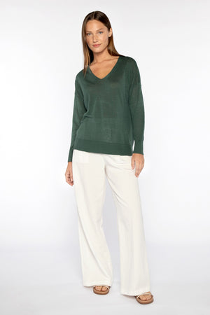 A woman smiles with one hand on her hip against a white background, wearing a KINROSS CASHMERE Easy Vee Pullover in Linen paired with white wide-leg pants, sandals, and her long hair down.