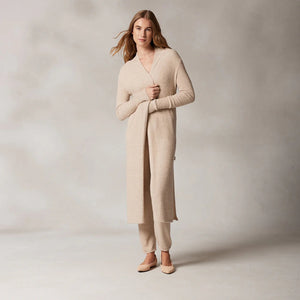 A woman with long hair wearing the KASHWERE - LONG CARDIGAN, part of a cozy beige lounge set from KASHWERE, crafted from Kash-ease yarn. The ensemble consists of a long-sleeved top, drawstring pants, and the matching long cardigan. She stands against a light grey background, looking confidently at the camera in her laid-back luxury outfit. Beige flat shoes complete the look.