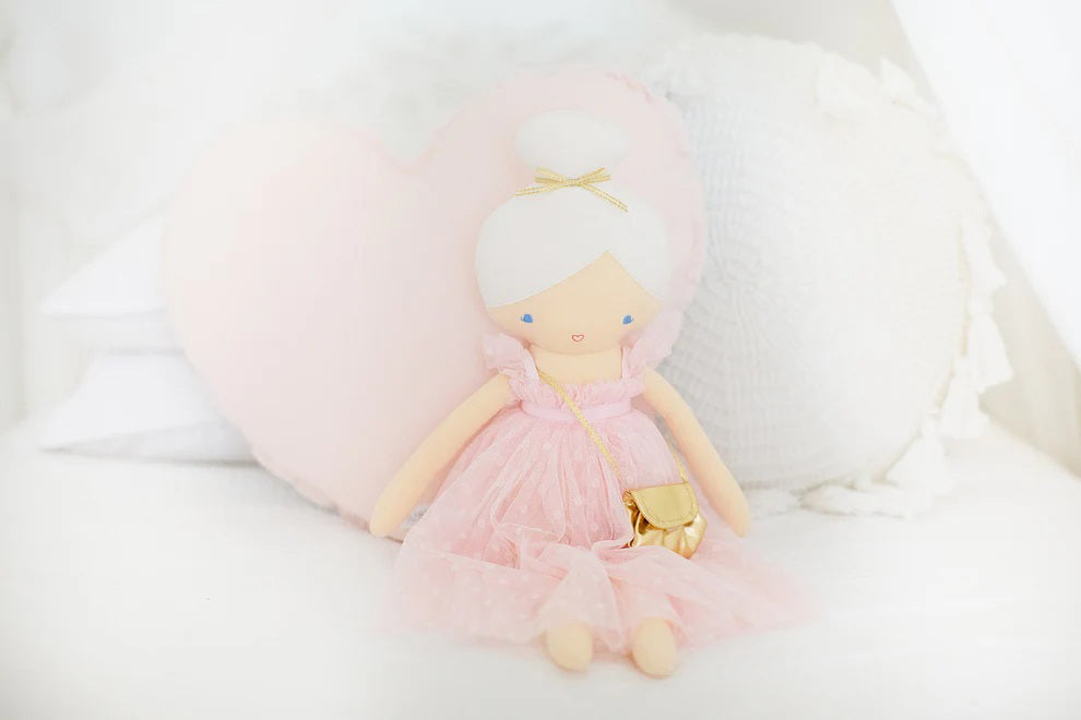 The ALIMROSE - CHARLOTTE DOLL PINK by ALIMROSE is a handcrafted doll with pale skin, dressed in a soft tulle light pink dress and carrying a golden purse. Her blonde hair is styled in a bun, and her small blue dot eyes coupled with a simple smile give her a charming expression.