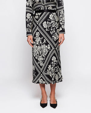 The MIRTO 1956 - BLACK AND WHITE PRINT MIDI SKIRT by MIRTO 1956 features a black A-line silhouette with an intricate white floral squares patchwork design set within diamond shapes. This skirt has a slight flare, providing a flowy appearance, and its symmetrical pattern offers a balanced and flattering chic look.