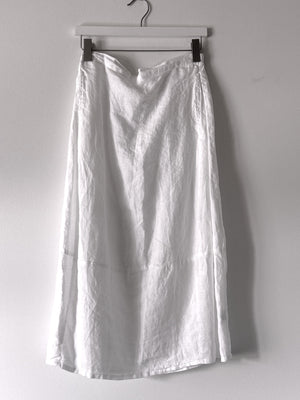 The CUT LOOSE - SIDE PLEAT SKIRT by CUT LOOSE hangs on a hanger against a plain grey background. The long white maxi skirt appears to be made of lightweight, slightly wrinkled linen fabric, with a simple design free of patterns or embellishments.