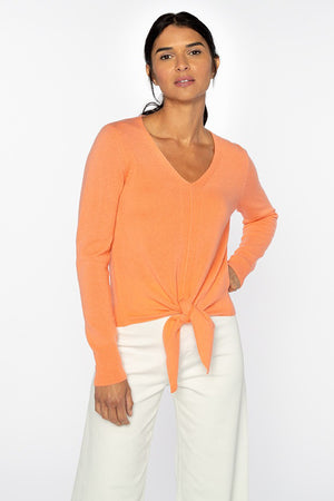 A person with long dark hair tied back stands confidently with one hand on their hip, wearing a KINROSS CASHMERE - TIE FRONT VEE PULLOVER by KINROSS. The 100% cashmere, 2ply, 9gg orange V-neck sweater features a stylish front tie detail and is paired elegantly with white wide-leg pants. The background is plain white.