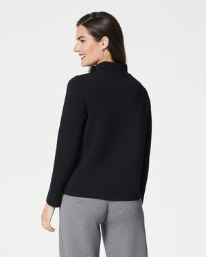 A person with long, dark hair smiles and poses with one hand in their pocket. They are wearing the SPANX - AIRESSENTIALS MOCK NECK TOP made from super-soft fabric, paired with matching dark green leggings against a light background.