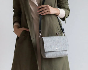 A person in a white paint-splattered shirt and tan pants holds the WOOLBERRY WOOL FELT CROSSBODY BAG, featuring a black strap, against a plain background.