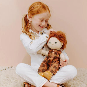 A Dinkum Doll from Olli Ella USA's Travel Togs collection, featuring dark hair and dressed in a denim jacket, mustard pants, and black shoes, stands on a wooden surface. Beside the doll is a teal See-Ya Suitcase with white trim and hardware. In the background, another larger teal suitcase is partially visible.