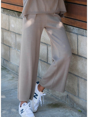 A person wearing MER SEA's SEAPORT KNIT PULL ON PANT in light brown, featuring a loose fit, wide legs, and side pockets, paired with white New Balance sneakers stands with one leg bent in front of a stone wall. The upper part of their body is partially visible, dressed in a matching light brown top.