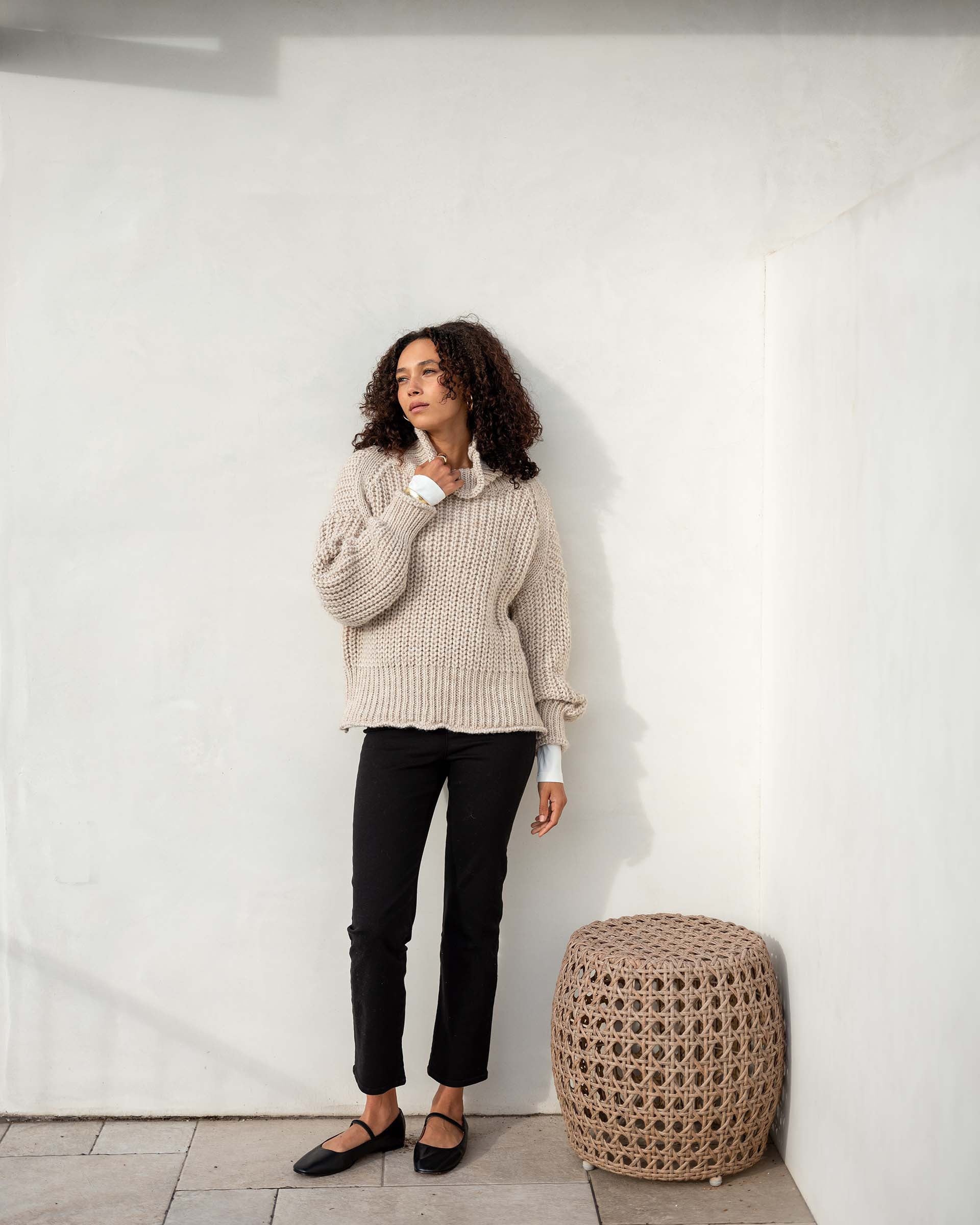 A woman with curly hair stands against a light-colored wall, wearing the MERSEA - BEACHSIDE TURTLENECK from MER SEA over a white shirt, black pants, and black flat shoes. Beside her is a woven cylindrical ottoman. She gazes thoughtfully to the side with a relaxed posture.