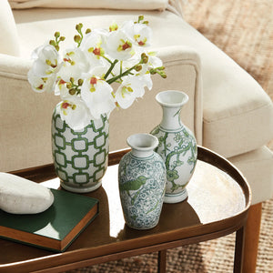 The NAPA HOME AND GARDEN - IMPERIAL BUD VASE, featuring green geometric and floral patterns, sits on a golden tray. Each handcrafted piece holds delicate peonies on a wooden table, beautifully illuminated by soft natural light filtering through the window.