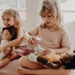 A close-up of a baby wearing OLLI ELLA USA DINKUM DOLL SHOES in Mallow Pink and white socks. The baby is seated on a soft, pale pink textured blanket, dressed in light brown clothing. Only the lower half of the baby's body is visible.