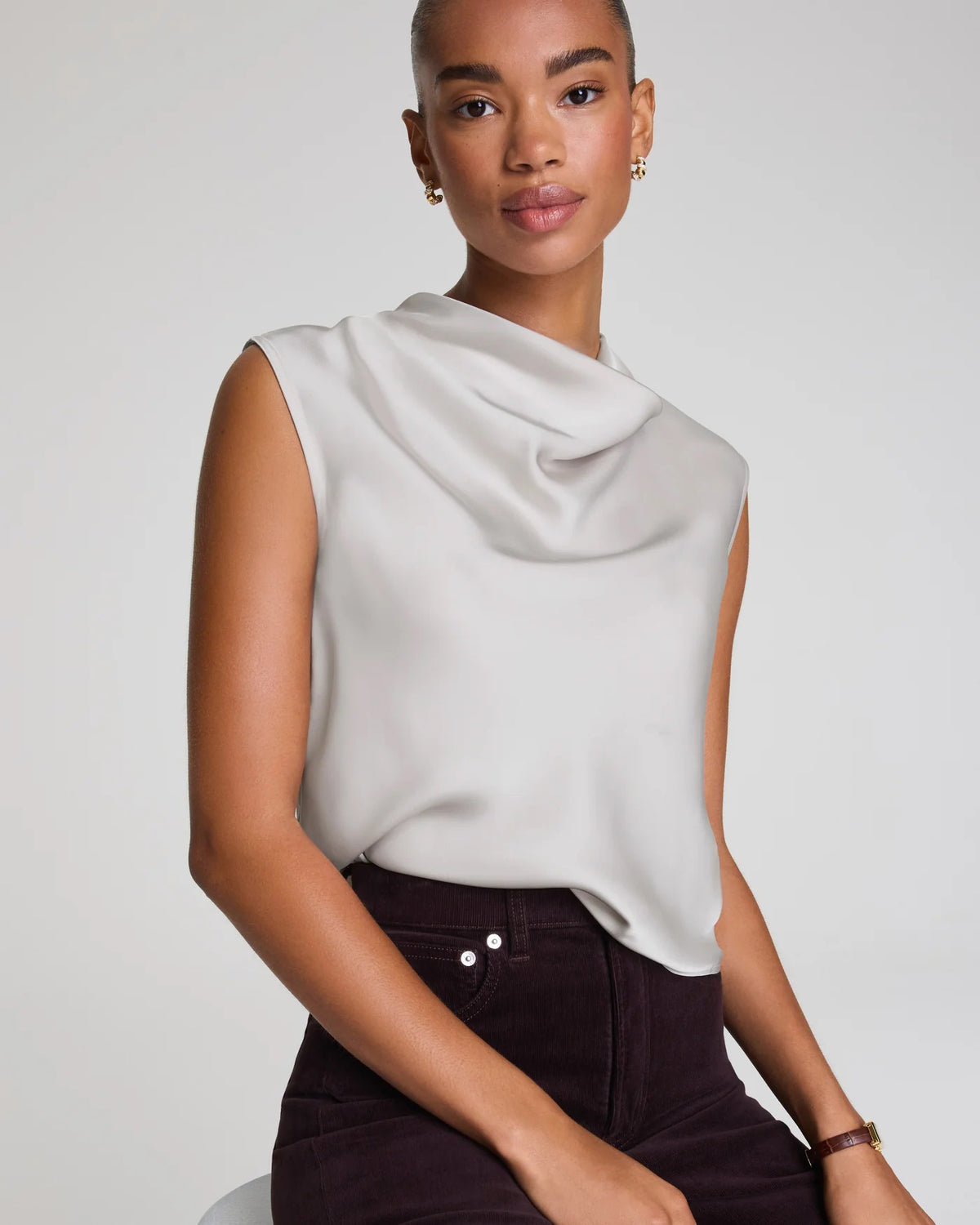 A woman wears the SPANX Silky Smooth Cowlneck Sleeveless Top in light gray with dark high-waisted pants, sitting on a stool against a plain background and looking at the camera with a neutral expression.