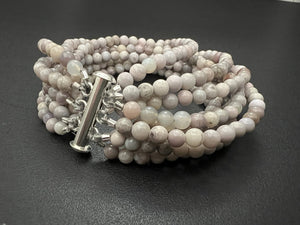 The ANN LIGHTFOOT - NINE STRAND SAKURA AGATE CUFF features smooth, round sakura agate beads in light grey and beige. Coiled with multi-strands, the bracelet has 4mm beads and is secured by a stainless steel clasp against a plain white backdrop.