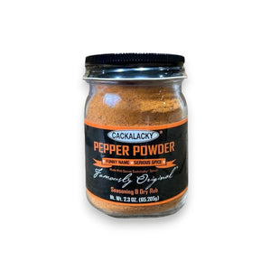 A glass jar of CACKALACKY - PEPPER POWDER RUB, made with fiery chile peppers and secret spices, is shown against a white background. The jar has a black lid and a black label with orange and white text stating "Cackalacky Pepper Powder," "Funny Name - Serious Spice," and "Famously Original." The jar contains 2.3 oz (65.2g).