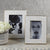 In a ZODAX MARMO MARBLE FRAME 5X7, two black and white photos are displayed on a patterned surface; one showcases a woman wearing a hat, while the other offers a close-up of her face. Accompanying the sophisticated photo arrangement is a decorative pot filled with white flowers, all set against a textured gray wall.