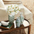 A cozy living room scene with a beige armchair in the background and a wooden side table in the foreground. The table holds three decorative vases—one featuring realistic stems of white NAPA HOME AND GARDEN's PHALAENOPSIS ORCHID STEM BUNDLE 6 orchids, and two adorned with intricate green patterns. A green book and a smooth stone also rest on the table, perfect to elevate your living space.