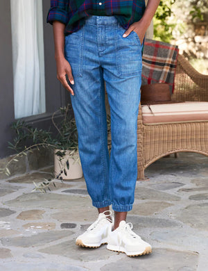 A person is standing on a stone patio with their back turned, wearing blue FRANK & EILEEN - JAMESON UTILITY JOGGER IN 1984 WASHED DENIM jeans with a high-rise fit, a plaid shirt with rolled-up sleeves, and white Veja sneakers. A wicker chair with a plaid blanket is visible in the background.