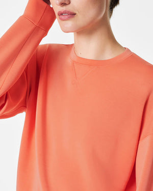 A woman stands confidently against a plain white background, wearing the SPANX Aireessentials Crew Neck Pullover and matching sweatpants made from luxurious comfort spacer fabric. She has her hair pulled back and her left hand is raised slightly. She smiles gently with her lips closed.