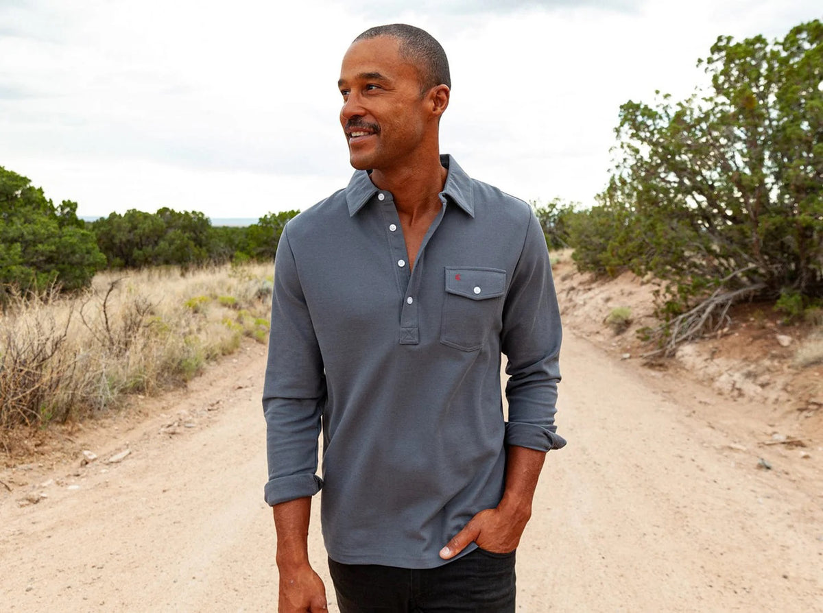 A man stands against a white background wearing the CRIQUET Long Sleeve Players Polo Shirt in Blue Steel with a chest pocket and dark jeans. His sleeves are rolled up, and his head is not visible.