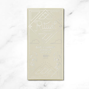 An opened brown paper package partially reveals a dark chocolate bar with intricate patterns. The package features a geometric design and the word "Ritual" written in a stylized font, followed by "One Hundred Percent Cacao Blend Chocolate Bar." The background is a white marble surface.