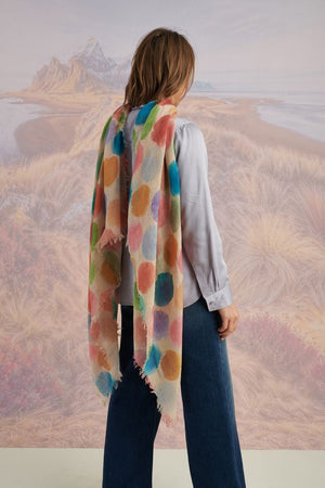 A woman with blonde hair displays the STORIATIPIC - BETTY WOOL SCARF, a large rectangular wool accessory with earth-toned polka dots, set against a scenic mountain landscape.