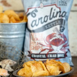 A small plate of kettle-cooked potato chips is shown in close-up, with a 5oz bag of Carolina Kettle - Crab Boil Chips by 1 In 6 Snacks in the background. Nearby, a metal container filled with chips and seashells captures the essence of a Carolina summer, celebrating southern tradition.