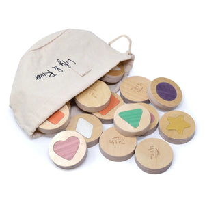 A young boy in a green shirt plays intently with the LITTLE MATCHABLES GAME SET by LILY & RIVER on a white table in a Montessori playroom. The room is adorned with various toys and baskets, and the boy appears focused on enhancing his fine motor skills by arranging and stacking the round wooden discs as part of this engaging memory game.