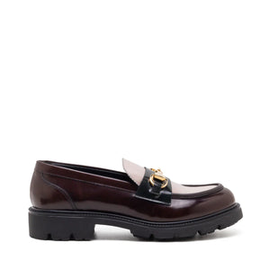 The MICHELE LOPRIORE - BRENDA TWO TONE LOAFER showcases a single dark brown leather design with a shiny finish. It features a black strap and gold-tone horsebit detailing across the top, complemented by a thick, chunky black sole for added durability. The shoe is set against a plain white background.