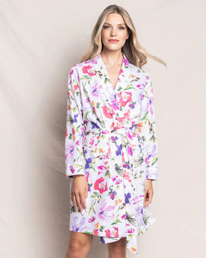 The PETITE PLUME - GARDENS OF GIVERNY ROBE by PETITE PLUME is a white, knee-length robe adorned with vibrant hazy florals in shades of pink, purple, orange, and green. This luxurious sleepwear features a shawl collar and a matching fabric belt tied around the waist, crafted from 100% finest quality cotton.