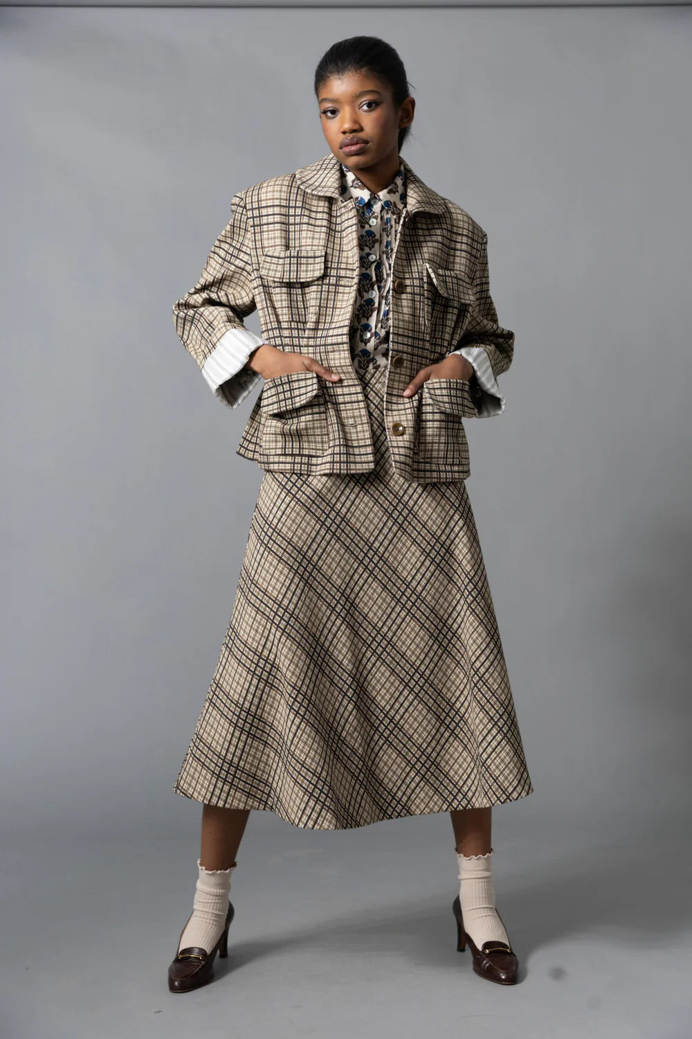 A woman stands sideways against a gray background, wearing the HOPE FOR FLOWERS - FOUR POCKET JACKET by HOPE FOR FLOWERS with a matching beige and brown plaid skirt, white socks, and dark brown heels. Her hair is pulled back as she looks slightly upward.