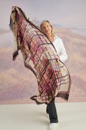 A woman in a blue shirt and jeans stands holding the STORIATIPIC - JULIE WOOL SCARF, a luxury plaid accessory in earthy tones. Behind her, mountains, beach, and grassy dunes create a scenic backdrop.