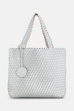 The WOVEN REVERSIBLE TOTE BAG by LINES OF DENMARK is a white tote featuring two shoulder straps and a circular faux-leather tag on one strap. This shopper showcases a textured, interlaced pattern and reversible design, providing stylish and elegant dual styling options. The image background is plain white.