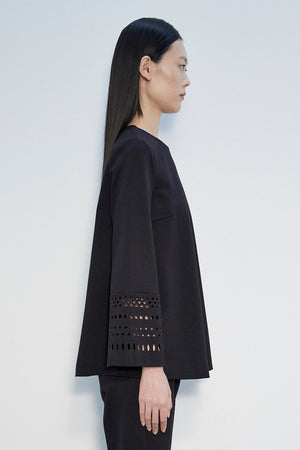 A person with long, straight, dark hair is standing with their back to the camera. They are wearing a stylish MEIMEIJ - LASERCUT SLEEVE SWING TOP with perforated sleeves near the cuffs, paired with matching black pants. The elegant ensemble exudes a unique charm, reflecting sophisticated designs made in Italy by MEIMEIJ.