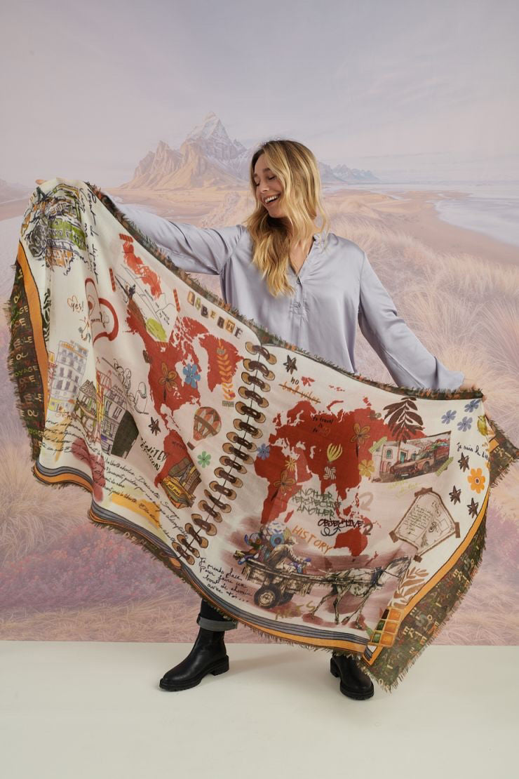 A woman with wavy blonde hair smiles while looking down. She wears a loose, light lavender blouse, dark pants, and the STORIATIPIC EUGENIE WOOL SCARF. Behind her is a scenic landscape of mountains, grass, and water.