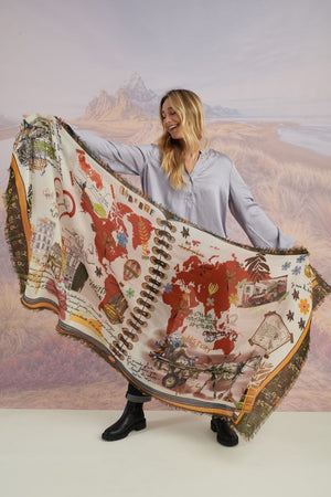 A woman with wavy blonde hair smiles while looking down. She wears a loose, light lavender blouse, dark pants, and the STORIATIPIC EUGENIE WOOL SCARF. Behind her is a scenic landscape of mountains, grass, and water.