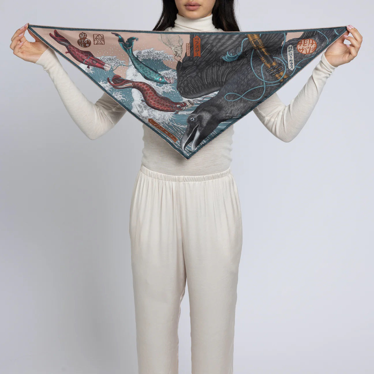 A person dressed in white is holding the SABINA SAVAGE - KOINOBORI KARASU TRIANGLE SILK SCARF from SABINA SAVAGE ENGLAND, which evokes the Edo period of Japan with its detailed marine illustrations of whales, a bear, and koinobori. The ocean-themed scarf is spread wide to display its vibrant design.