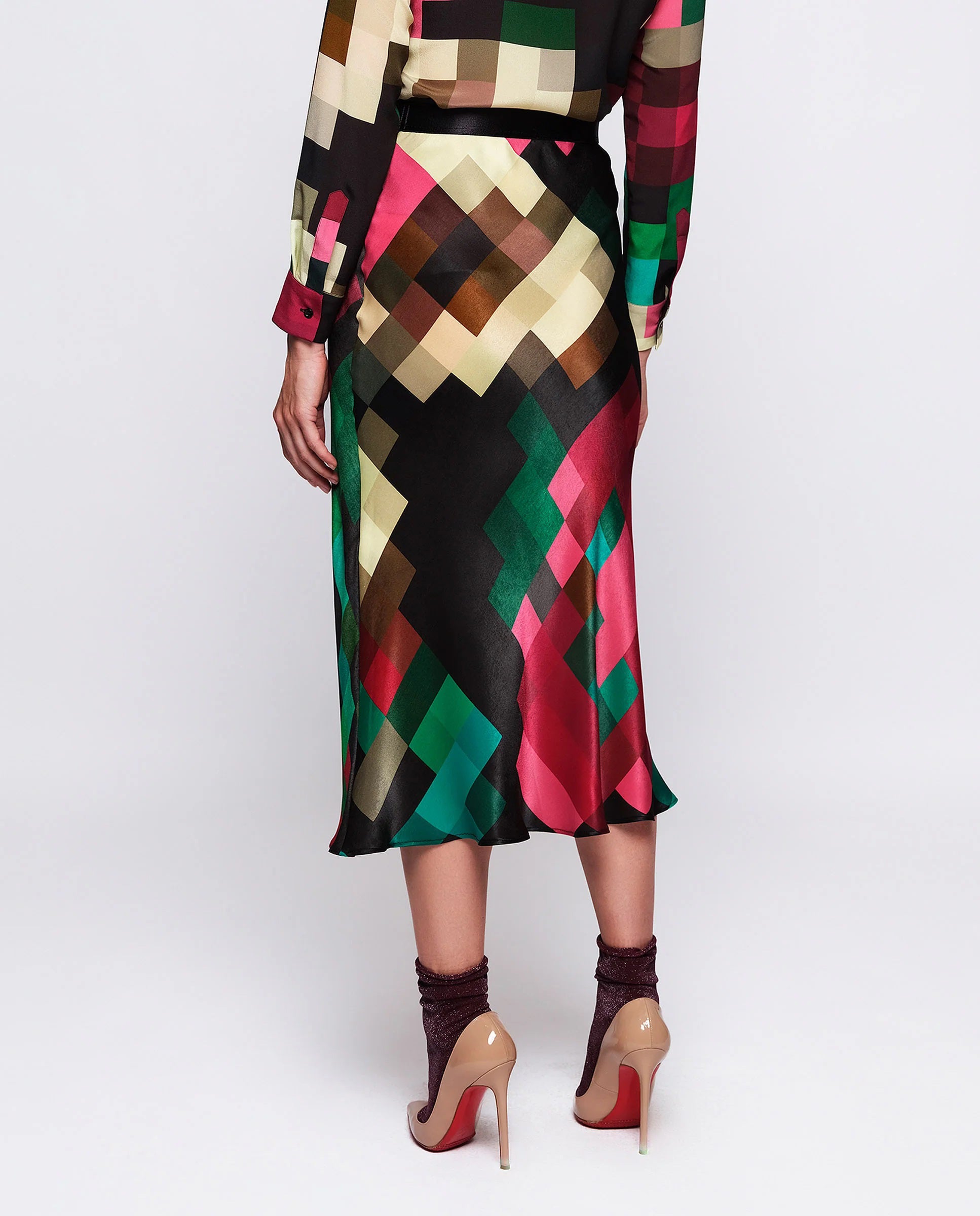 A person is wearing the MIRTO 1956 MULTICOLOR PIXEL PLAID MIDI SKIRT, which boasts a multi-colored, geometric pattern with an elastic satin waistband. They have paired it with a matching long-sleeve top, glittery ankle socks, and high-heeled shoes. The skirt features a vibrant mix of red, green, black, and beige colors that evoke a pixel print design. The background is plain white.