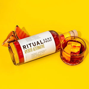 A tall, clear bottle with a yellow cap contains a red liquid. The label reads "RITUAL ZERO PROOF- APERITIF ALTERNATIVE" in large, modern font, along with additional small text detailing the product. The design is minimalistic, featuring white, yellow, and black colors.