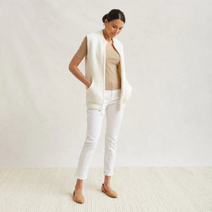 A woman stands against a light gray background wearing the KASHWERE - UNISEX VEST by KASHWERE, a sleeveless, zip-up cream-colored knit vest over a short-sleeve beige top. She has her hands in the cozy layering pockets and is dressed in white pants. Her dark hair is pulled back as she looks at the camera with a slight smile.