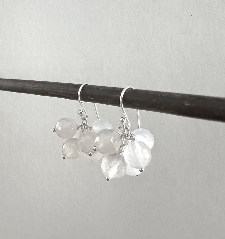 The ANN LIGHTFOOT MILKY AGATE CLUSTER EARRINGS, designed by ANN LIGHTFOOT, dangle from a slender, dark rod against an unadorned backdrop. Each earring showcases numerous translucent, faceted beads clustered together and suspended from a minimalist sterling silver hook enhanced with milky agate stones.