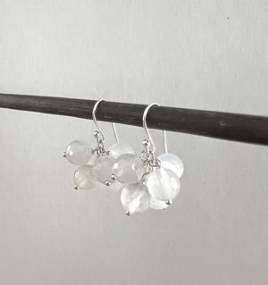 The ANN LIGHTFOOT MILKY AGATE CLUSTER EARRINGS, designed by ANN LIGHTFOOT, dangle from a slender, dark rod against an unadorned backdrop. Each earring showcases numerous translucent, faceted beads clustered together and suspended from a minimalist sterling silver hook enhanced with milky agate stones.